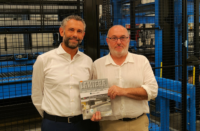 Two men in white shirts stand in front of industrial machinery, holding a magazine.