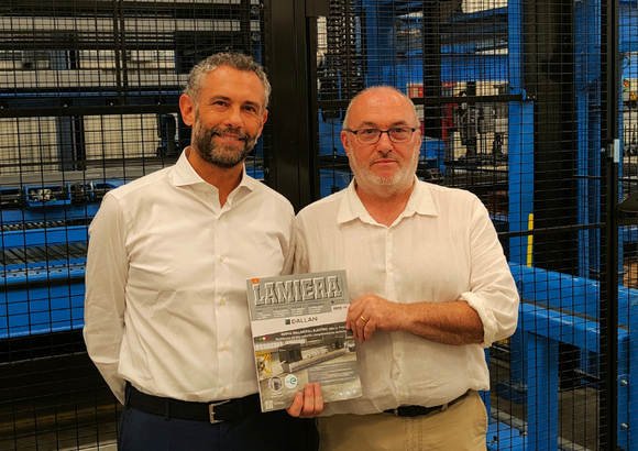 Two men in white shirts stand in front of industrial machinery, holding a magazine.