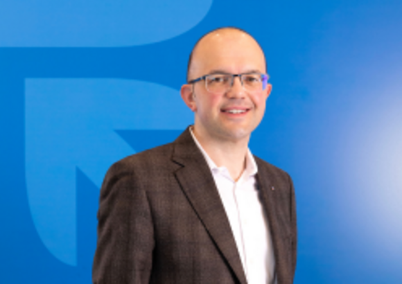 Smiling man in glasses and blazer against a blue background with a large logo