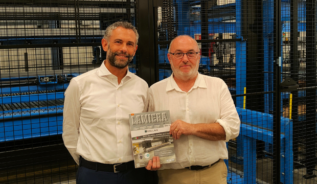 Two men in white shirts stand in front of industrial machinery, holding a magazine.